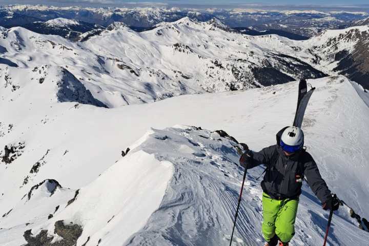 Skier climbing a snowy mountain ridge with bright blue sky and distant peaks in background.