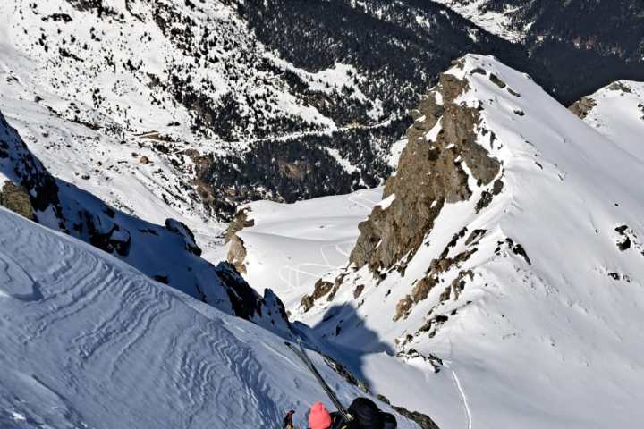 Person climbing snowy mountain with ski gear, rugged snowy peaks in background, clear blue sky.
