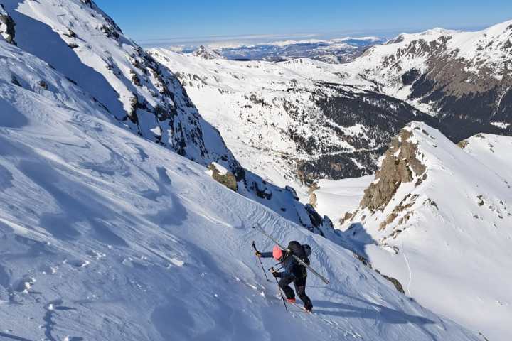 Person hiking up a snowy mountain slope with ski poles, under a clear blue sky.