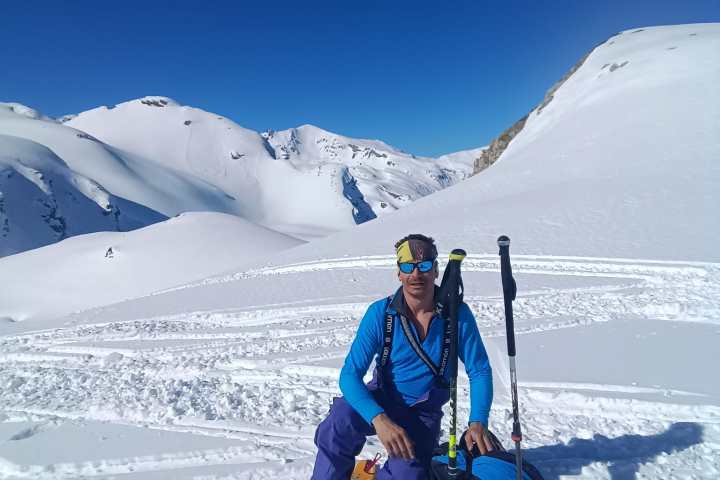 Person in skiing gear kneeling on snowy mountain slope against clear blue sky.