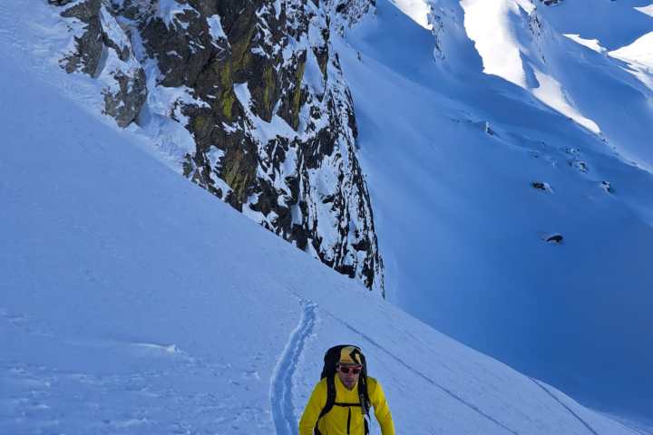 Skier in yellow jacket climbs snowy mountain trail beside steep rock face under clear blue sky.