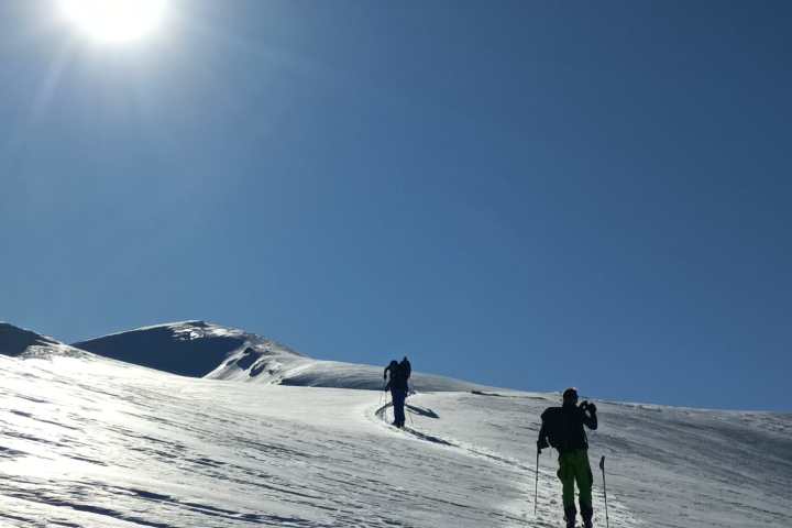 Skiers ascending snowy slope under bright sun in clear blue sky.