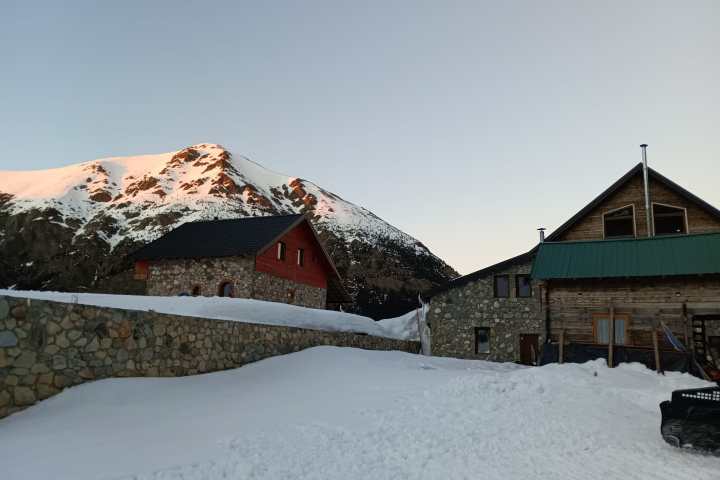 Snow-covered mountain and stone cabin under a clear sky at sunrise.