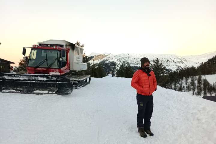 Person in a red jacket stands on a snowy mountain path near a red snow groomer.