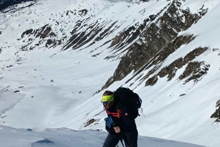 Person in ski gear climbing snowy mountain with ski poles, surrounded by snow-covered peaks under a clear blue sky.