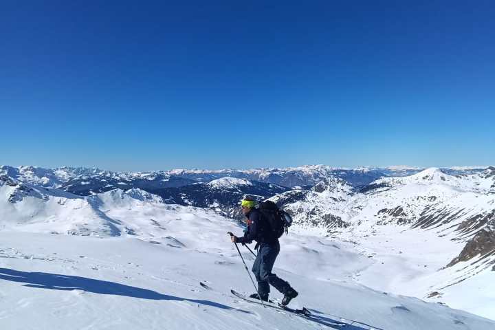 Skier climbing a snowy slope with mountains in the background under a clear blue sky.