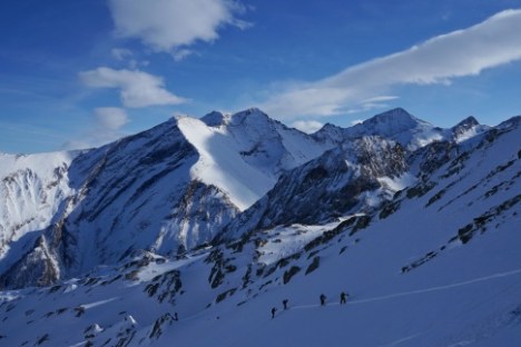 Skiers climbing snow-covered mountain slope with rugged peaks under a blue sky.