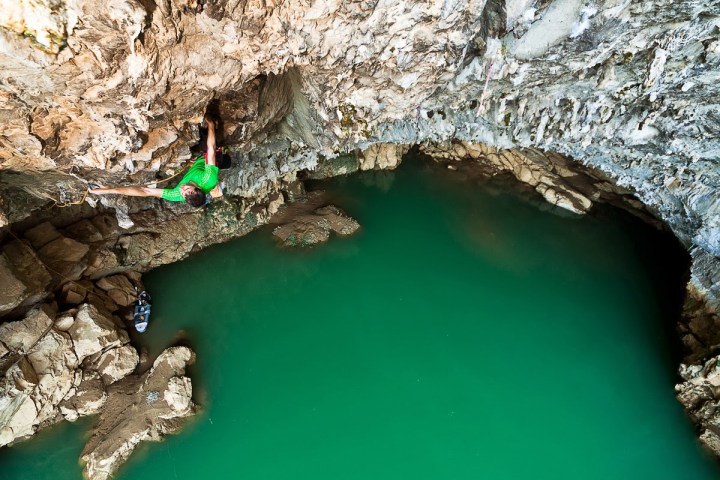 Rock climber in green shirt scaling a cave wall above turquoise water.