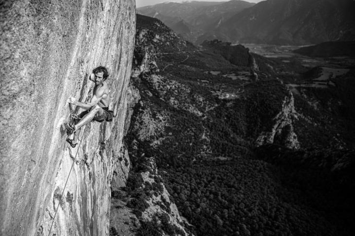 Shirtless man rock climbing a steep cliff with vast mountain view in black and white.