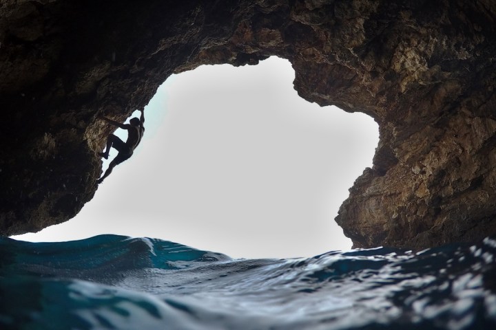 Silhouette of a person rock climbing inside a cave near water.