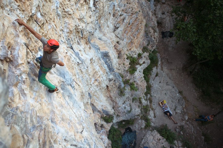 Climber with red helmet on a steep rock face, belayer resting below.