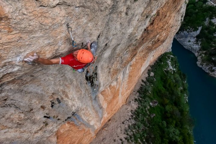 Person in orange helmet rock climbing a steep cliff above a river.