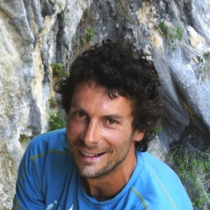 Smiling man with curly hair in blue shirt near a rocky background.