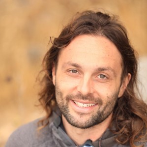 Smiling man with long brown hair and beard, wearing a gray jacket.