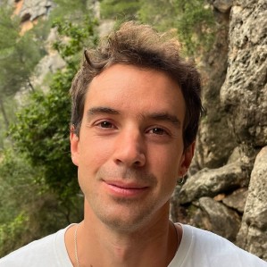 Person with short hair smiling outdoors, surrounded by trees and rocks.
